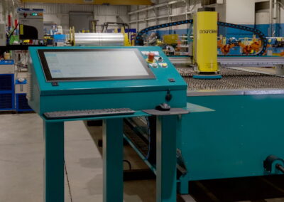 teal industrial control console with touchscreen and keyboard beside a teal CNC/press machine in a factory setting.