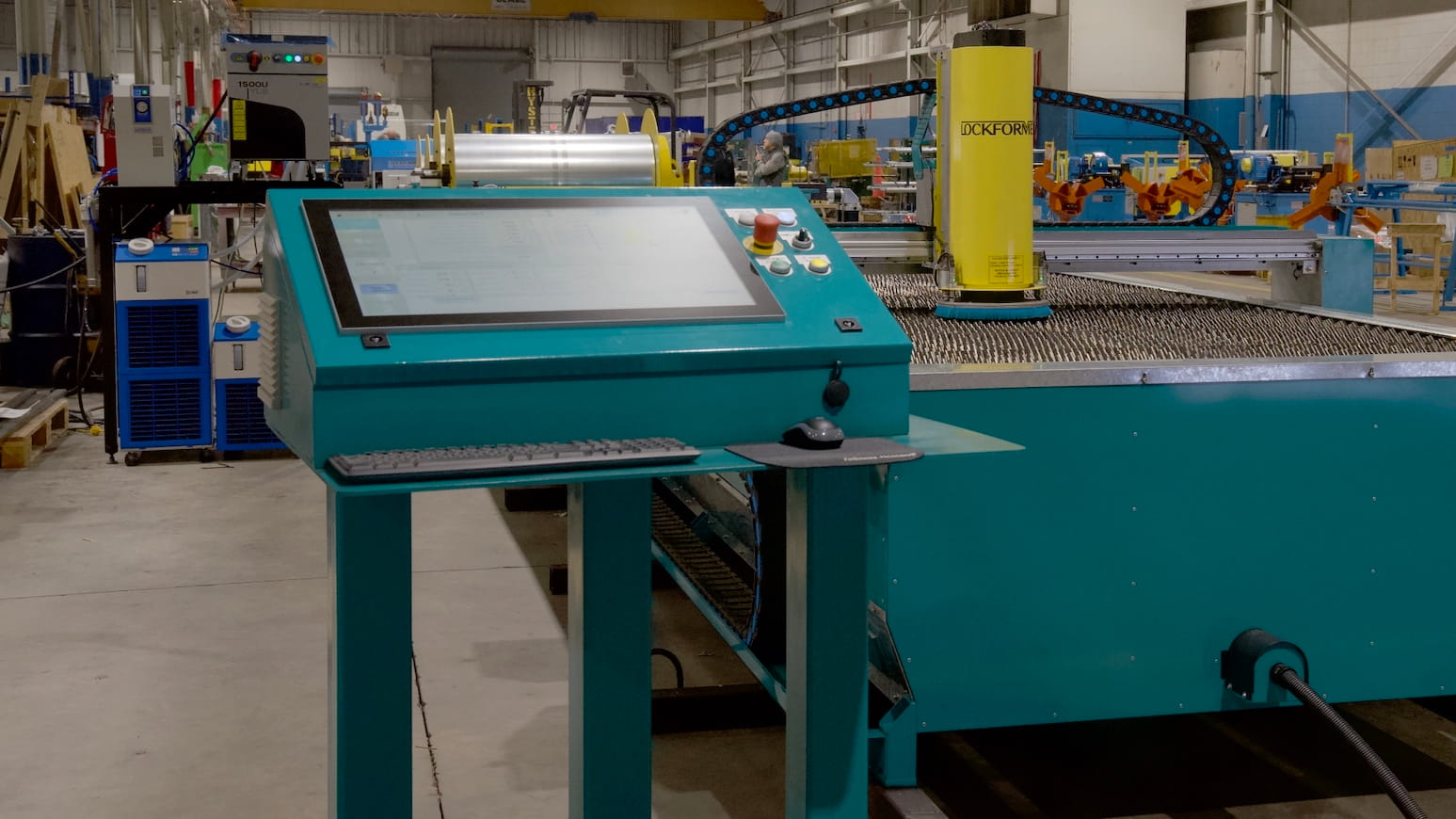 Man in black shirt operates teal industrial rollers on a busy factory floor with yellow safety lines and scattered equipment in the background.