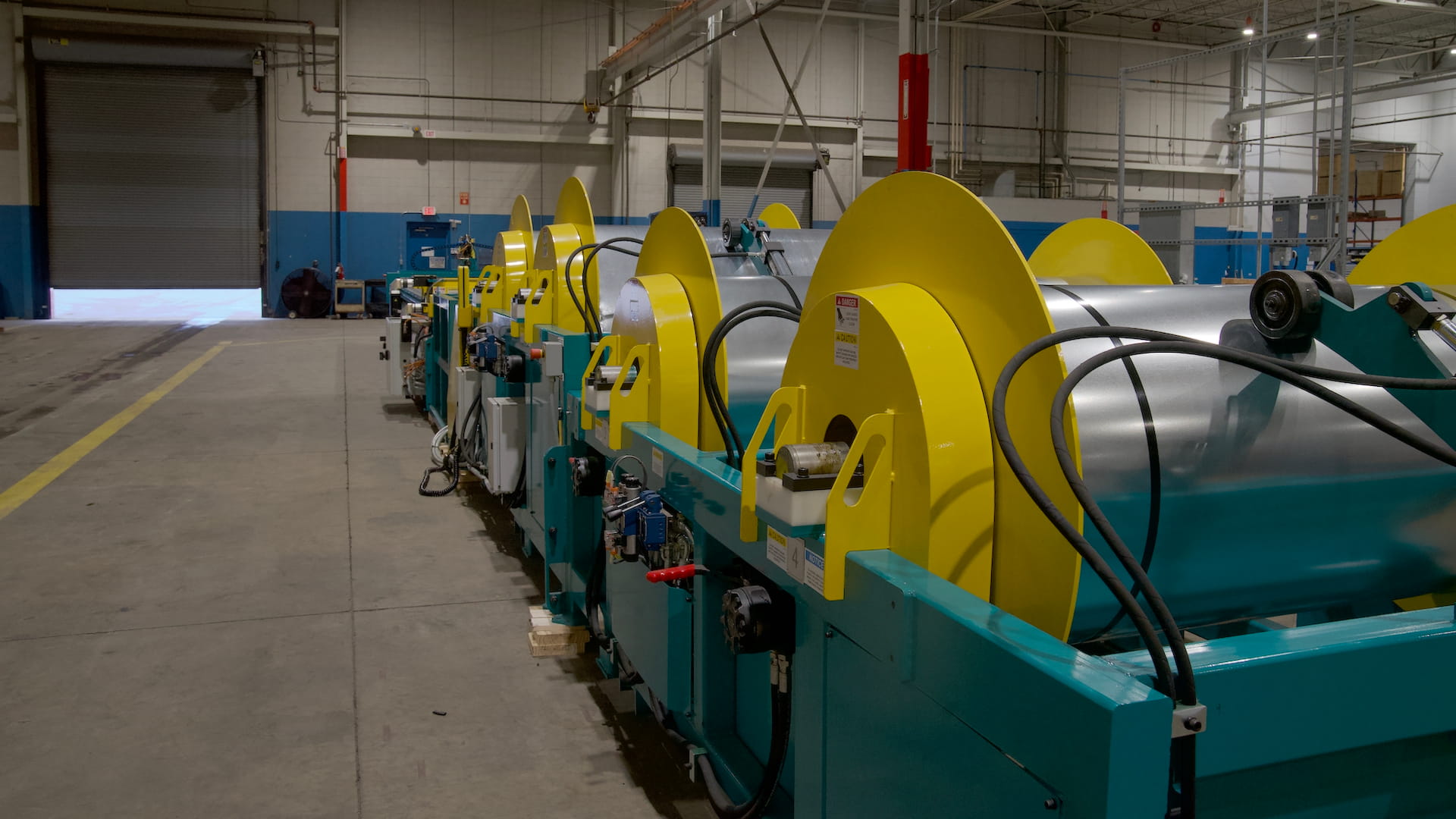 Industrial manufacturing line with yellow circular rollers and metal coils on teal frames inside a factory.