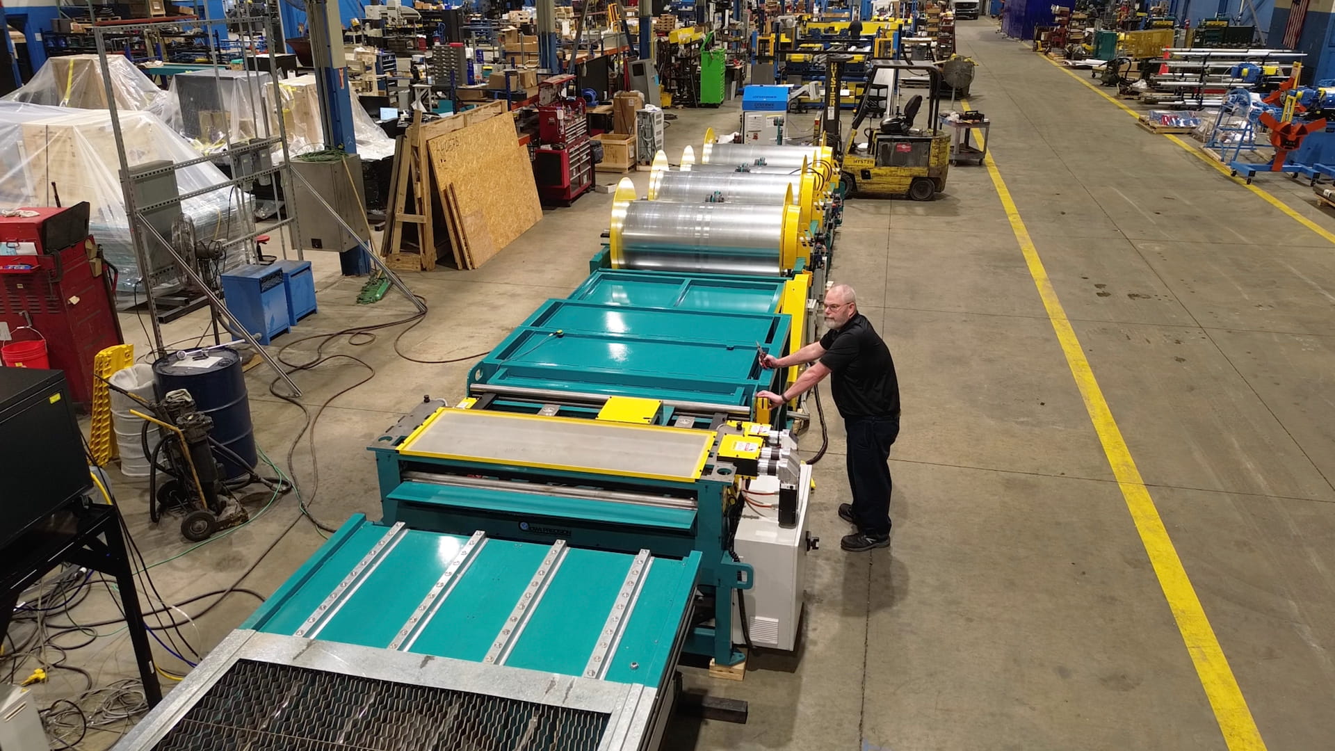 Man in black shirt operates teal industrial rollers on a busy factory floor with yellow safety lines and scattered equipment in the background.