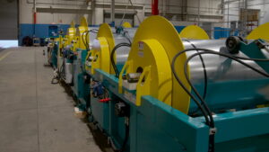 Industrial production line with large yellow spool rollers mounted on a teal frame inside a factory.
