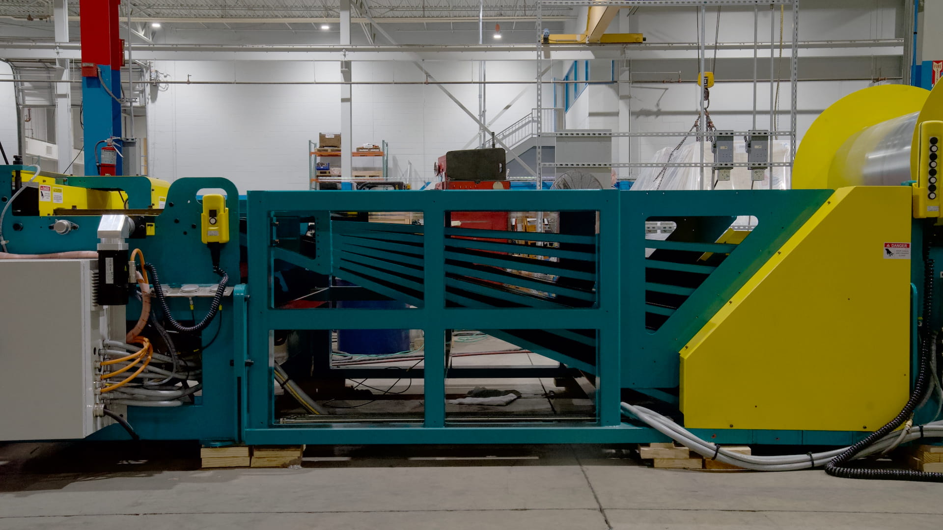 Industrial manufacturing machine with a teal frame and bright yellow safety cover in a factory, with cables and metal framework surrounding it.