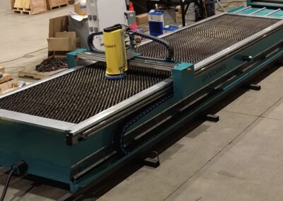 Industrial fabricating machine with a yellow component mounted over a metal mesh table in a shop setting, cables and pallets in the background