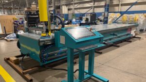 Teal-colored manufacturing machine with a long conveyor belt in a factory setting; control panel with keyboard and buttons in the foreground.