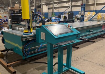 Teal-colored manufacturing machine with a long conveyor belt in a factory setting; control panel with keyboard and buttons in the foreground.