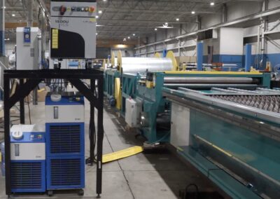 Industrial factory scene with a long sheet metal roller and green/blue conveyor system, yellow safety guards, and control cabinets under bright overhead lights.