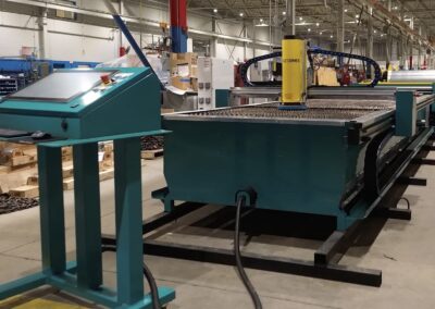 Factory floor with a large teal CNC-style machine and a yellow vertical actuator; control panel in the foreground.