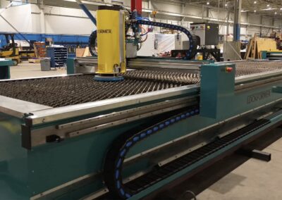 Teal Lockformer production line with a yellow vertical control unit and blue cable chain along a metal conveyor in a large factory floor