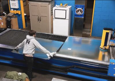 Person in a gray hoodie wipes a long blue industrial machine in a workshop with blue walls and cabinets nearby.