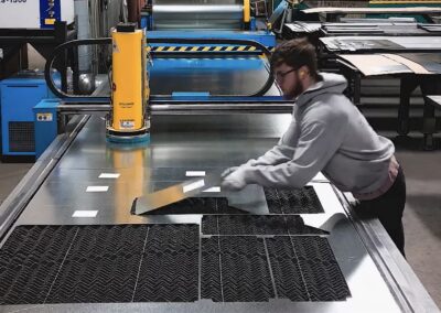 Worker in hoodie cutting sheet metal on a large industrial cutting table in a workshop, with a yellow machine and stacked metal sheets in the background.