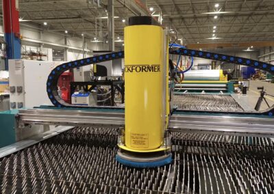 Industrial manufacturing floor with a bright yellow vertical machine at the center, surrounded by a blue-roller chain system and a metal slat conveyor bed.