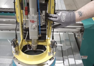 Gloved hand operates a vertical spindle on a yellow machining frame in a factory setting, with metal rails and a green enclosure nearby.