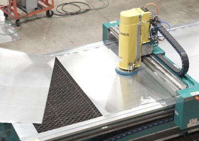 Worker guides a shiny metal sheet into a yellow vertical cutting head on an industrial sheet-metal cutter; a turquoise machine bed, a black mesh scrap area, and coiled hoses visible in the background.
