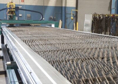 Long metal conveyor/roller table extending into a factory, with industrial equipment and chain racks in the background